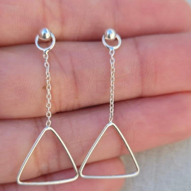 Silver triangle earrings held between fingers against a neutral background