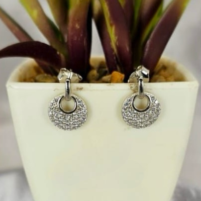 A pair of sterling silver pavé studs earrings with cubic zirconia, displayed in a white pot with purple and green plants in the background.