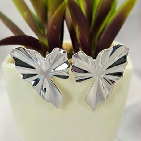 A pair of silver leaf-shaped stud earrings displayed on a white surface, with a green plant in the background.