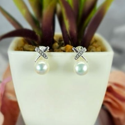 A pair of freshwater pearl stud earrings with a bling cross top design, displayed in a white vase with green leaves.