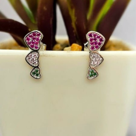A pair of heart-shaped stud earrings with a gold and silver color scheme, featuring cubic zirconia, displayed in front of a potted plant.