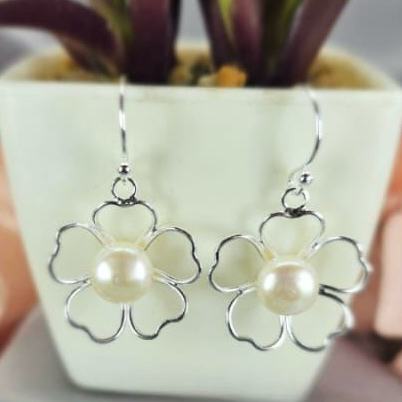 A pair of sterling silver drop earrings featuring a floral design with freshwater pearl centers, displayed in front of a potted plant.