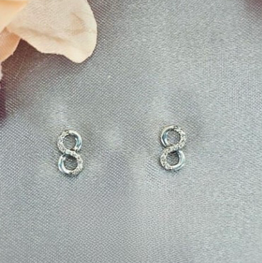 A pair of sterling silver infinity stud earrings with pavé cubic zirconia, displayed on a grey surface with a flower in the background.