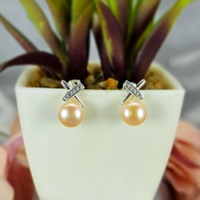 A pair of gold-colored freshwater pearl earrings with a bling cross top design, displayed in a white pot with green leaves.