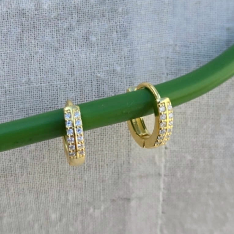 Gold hoop earrings with clear stones on a green stick against a gray background