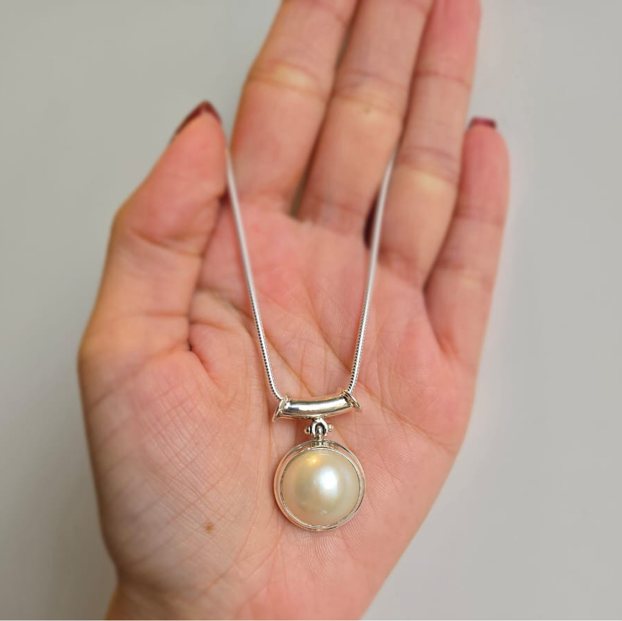 An 18mm white mabe pearl pendant attached to a sterling silver necklace, held in a hand against a neutral background.