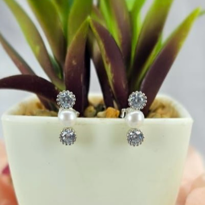 A pair of sterling silver stud earrings featuring a central freshwater pearl with two surrounding cubic zirconia crystals, displayed in a white vase with a green plant.