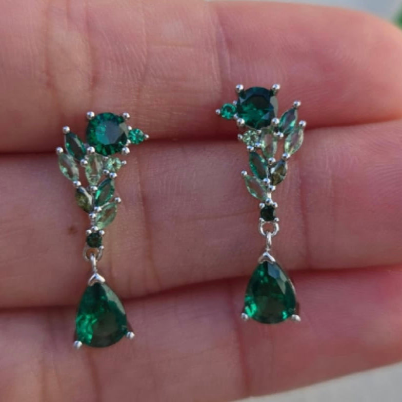 Green gemstone earrings held between fingers against a blurred background