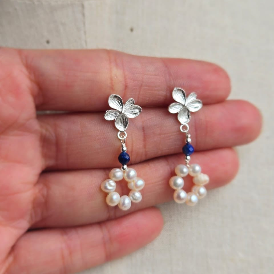 Silver earrings with floral designs and pearl beads held in a hand against a neutral background