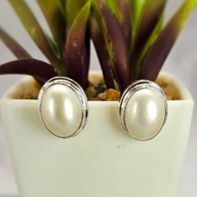 A pair of oval white Mabe pearl earrings set in sterling silver, displayed in front of a potted plant.