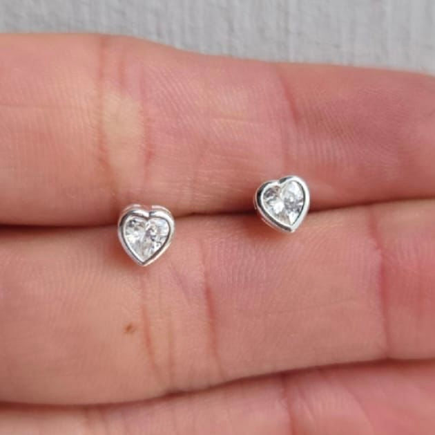 Heart-shaped silver earrings with clear gemstones on a hand.