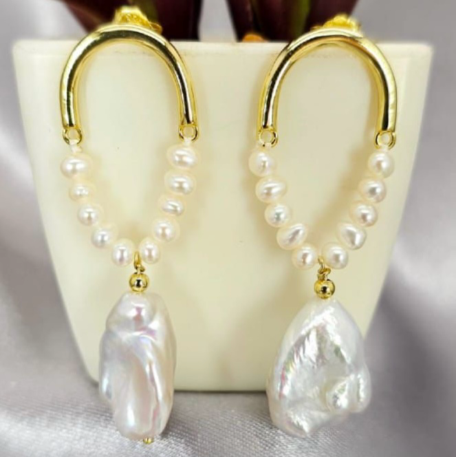 Gold statement earrings with pearls on end, displayed on a surface with a white vase and green foliage in the background.