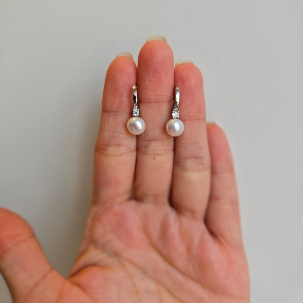 A pair of silver pearl drop earrings with cubic zirconia stones, displayed on a person's hand.