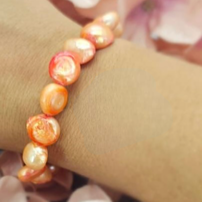 A peach-colored freshwater pearl bracelet displayed on a person's wrist with a background of pink petals.