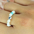 A person's finger wearing a ring adorned with tiny freshwater pearls in a white color, arranged on an elastic band.