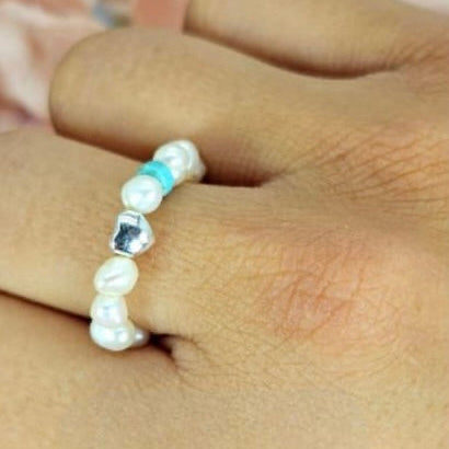 A person's finger wearing a ring adorned with tiny freshwater pearls in a white color, arranged on an elastic band.