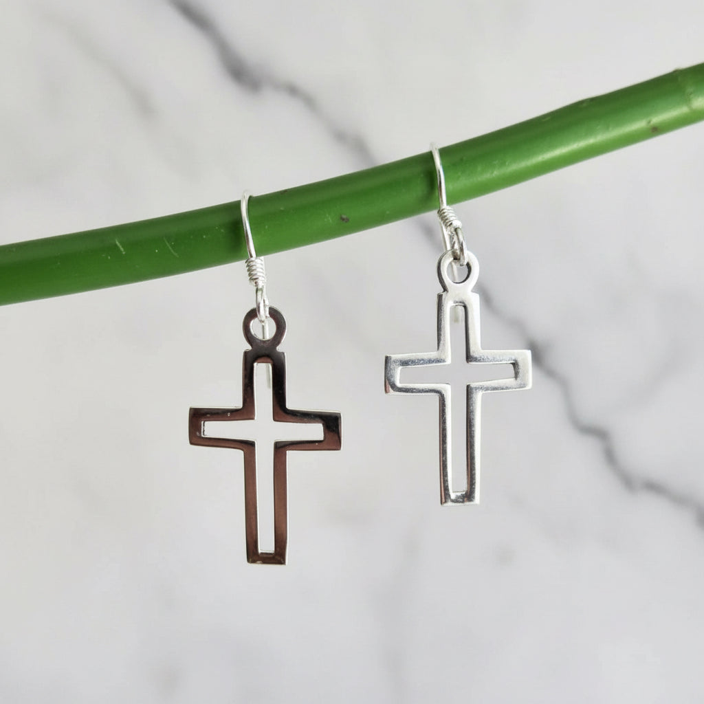 Silver cross-shaped earrings hanging from a green stick on a textured fabric background