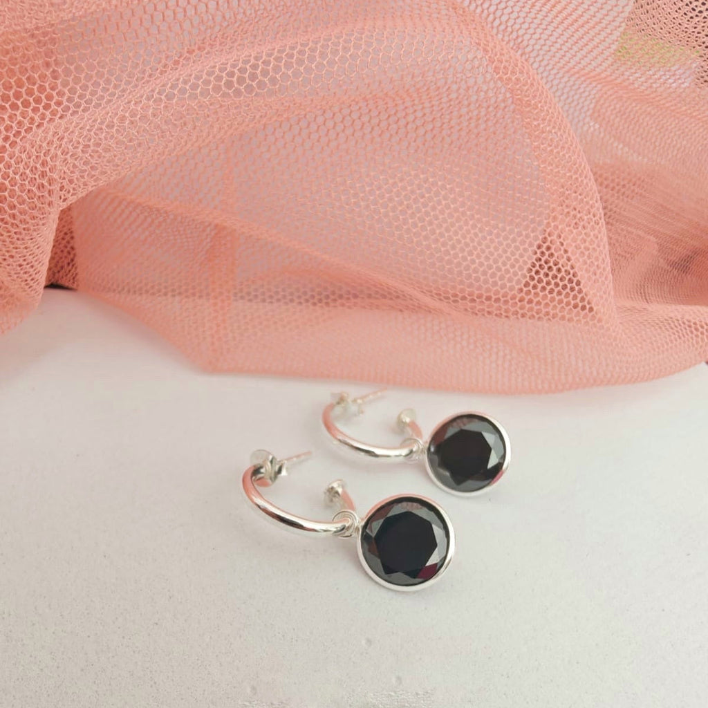Silver hoop earrings with black stones on a white surface with pink fabric in the background