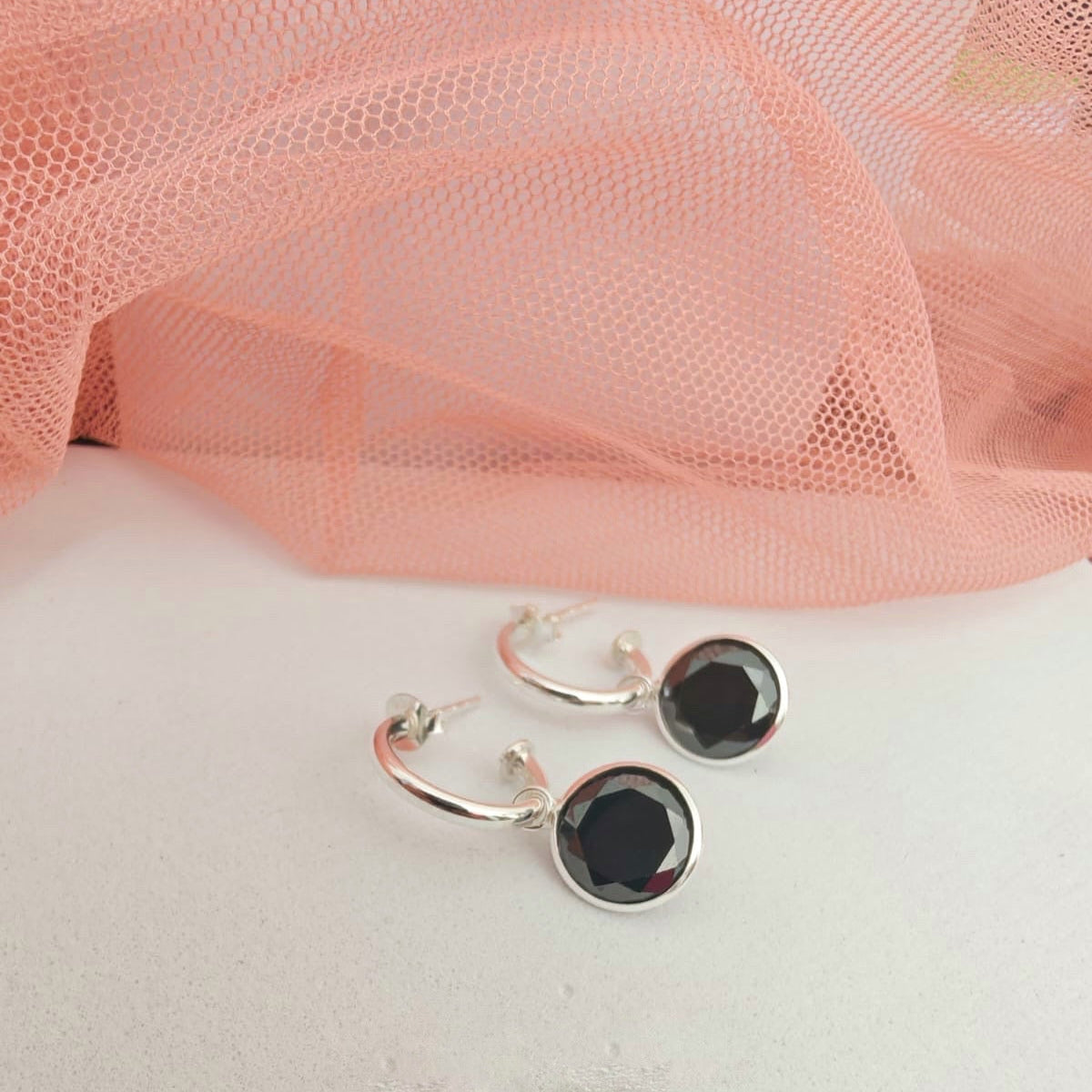 Silver hoop earrings with black stones on a white surface with pink fabric in the background