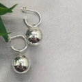A pair of sterling silver hoop earrings with a 16mm ball charm dangle, displayed on a grey background with a green leaf.