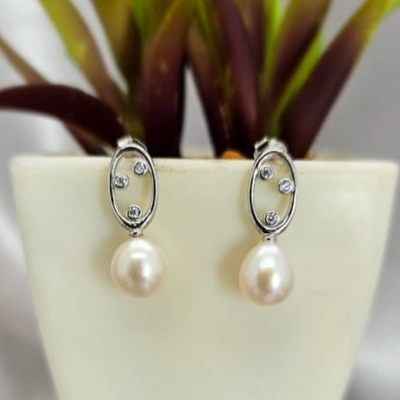 A pair of sterling silver earrings with freshwater pearl drops, displayed in a white vase with a green plant in the background.