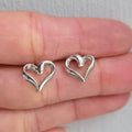 Silver heart-shaped earrings held between fingers on a neutral background