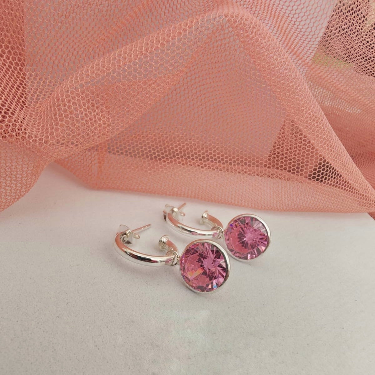 Silver hoop earrings with pink gemstones on a white surface with pink fabric in the background