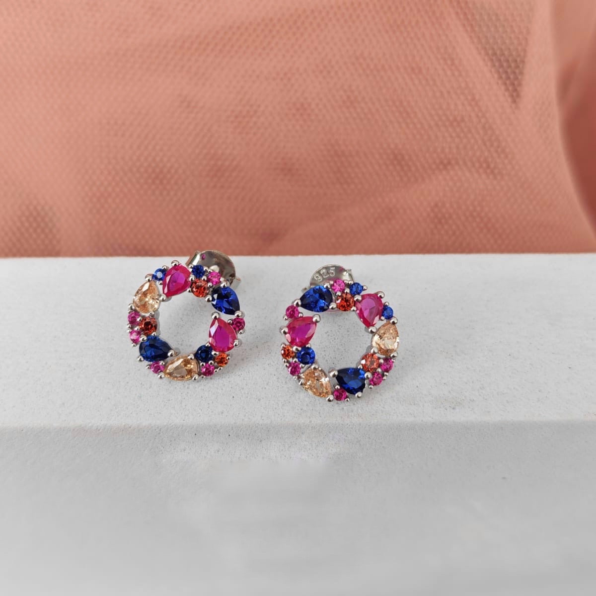 Multicolored circular earrings on a white surface with a pink fabric background