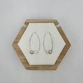 A pair of modern hook drop earrings with freshwater pearls displayed on a wooden hexagonal stand against a white background.