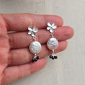 Silver earrings with pearl and black bead details held between fingers on a neutral background