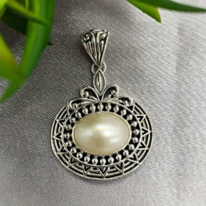An oval-shaped sterling silver pendant featuring a central Mabe pearl, surrounded by decorative elements in a geometric pattern, placed on a textured surface.