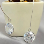 A pair of sterling silver drop earrings with round cubic zirconia crystals, displayed in front of a potted plant.