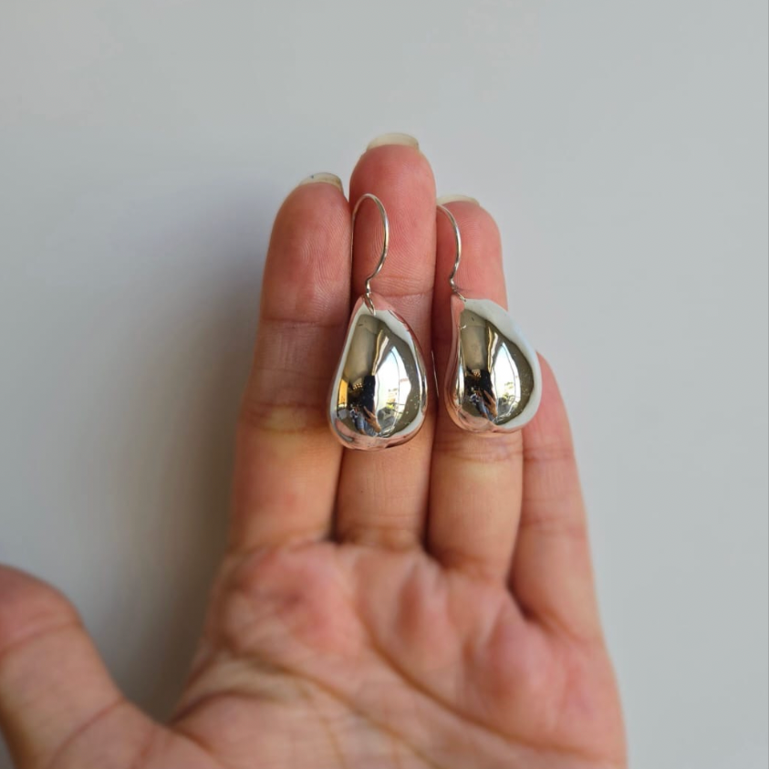 A pair of large teardrop-shaped earrings made from sterling silver, displayed on a person's hand.