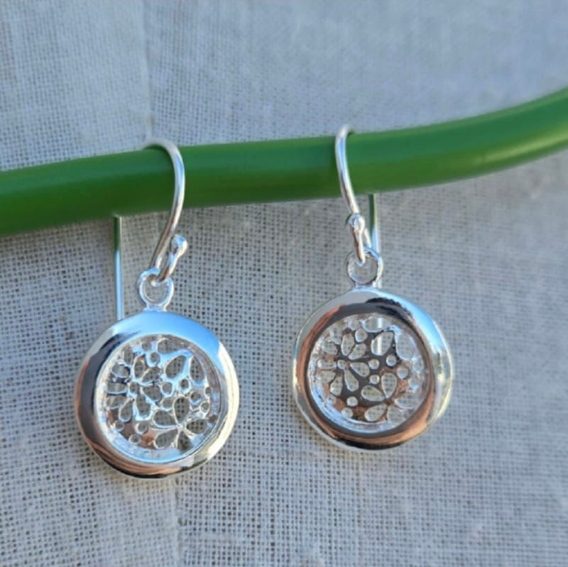 Silver earrings with intricate designs hanging on a green rod against a beige fabric background