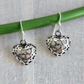 Silver heart-shaped earrings on a green stick against a textured fabric background