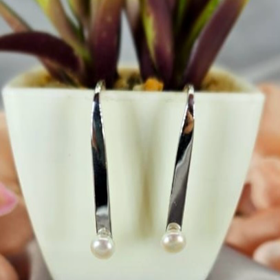 A pair of modern silver bar earrings with freshwater pearls on ends, displayed in a white pot with green plant leaves.
