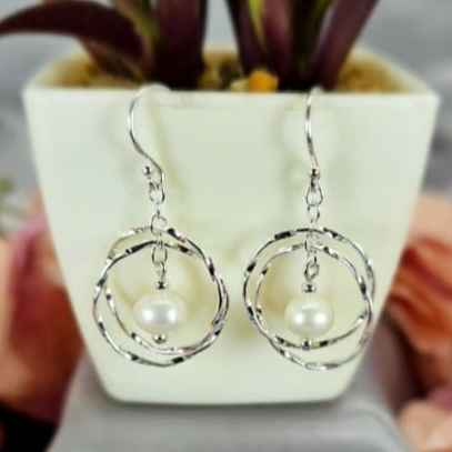 A pair of silver drop earrings with freshwater pearl accents, displayed on a white surface next to a potted plant.