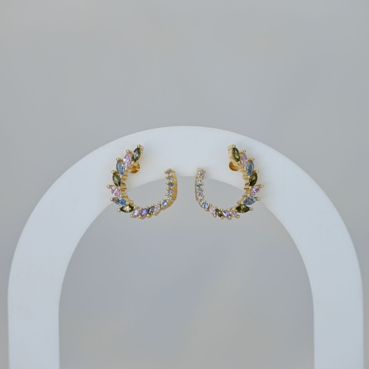 Pair of gold hoop earrings with colorful stones on a white stand against a light gray background