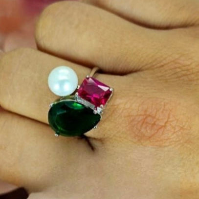 An image displaying a sterling silver ring with an emerald green teardrop, a red rectangular cubic zirconia, and a large freshwater pearl on a person's hand.