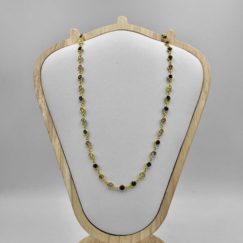 A gold necklace with green and black cubic zirconia stones displayed on a white cushion atop a wooden stand.