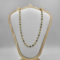 A gold necklace with green and black cubic zirconia stones displayed on a white cushion atop a wooden stand.