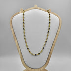 A gold necklace with green and black cubic zirconia stones displayed on a white cushion atop a wooden stand.