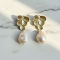 A pair of gold-plated earrings designed with flower petals and pearl drops, displayed in a white vase with green leaves.