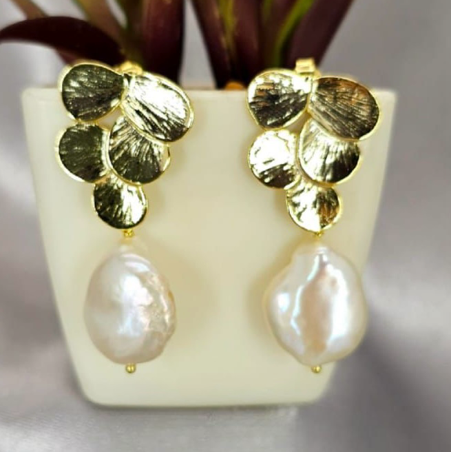 A pair of gold-plated earrings designed with flower petals and pearl drops, displayed in a white vase with green leaves.