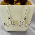 A pair of sterling silver hanging dove earrings displayed in front of a potted plant.