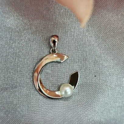 A sterling silver pendant in the shape of a C with a freshwater pearl attached to it, displayed on a grey fabric surface.