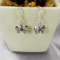 Sterling silver drop earrings with a butterfly design, displayed in a white vase with a green plant background.