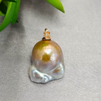 A 20 mm rough pearl pendant with a gold hook on a grey surface, partially covered by a green leaf.