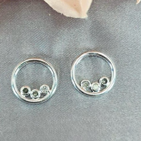 A pair of sterling silver round stud earrings with three round cubic zirconia stones each, displayed against a fabric background.