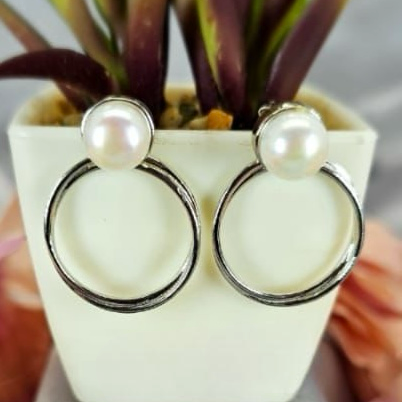 A pair of sterling silver hoop earrings with freshwater pearls, displayed in a white vase with a green plant and pink flowers around.